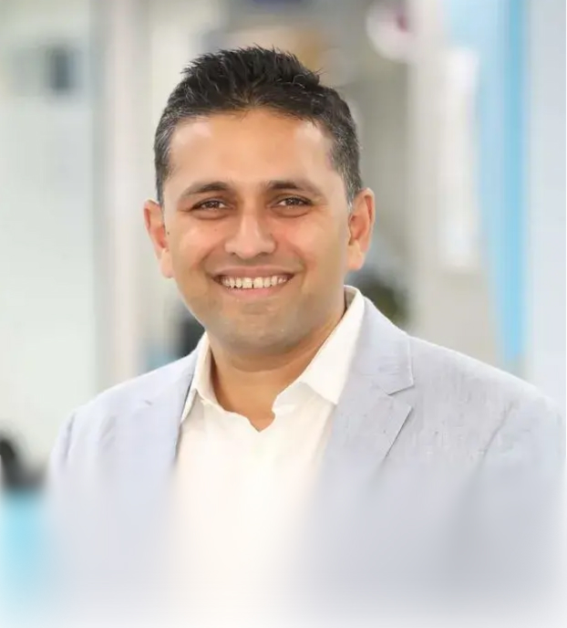 Smiling man with short dark hair wearing a light gray blazer and white shirt in an indoor setting.