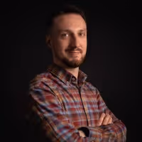 Smiling man with beard and mustache wearing a plaid shirt, standing with arms crossed against a dark background.