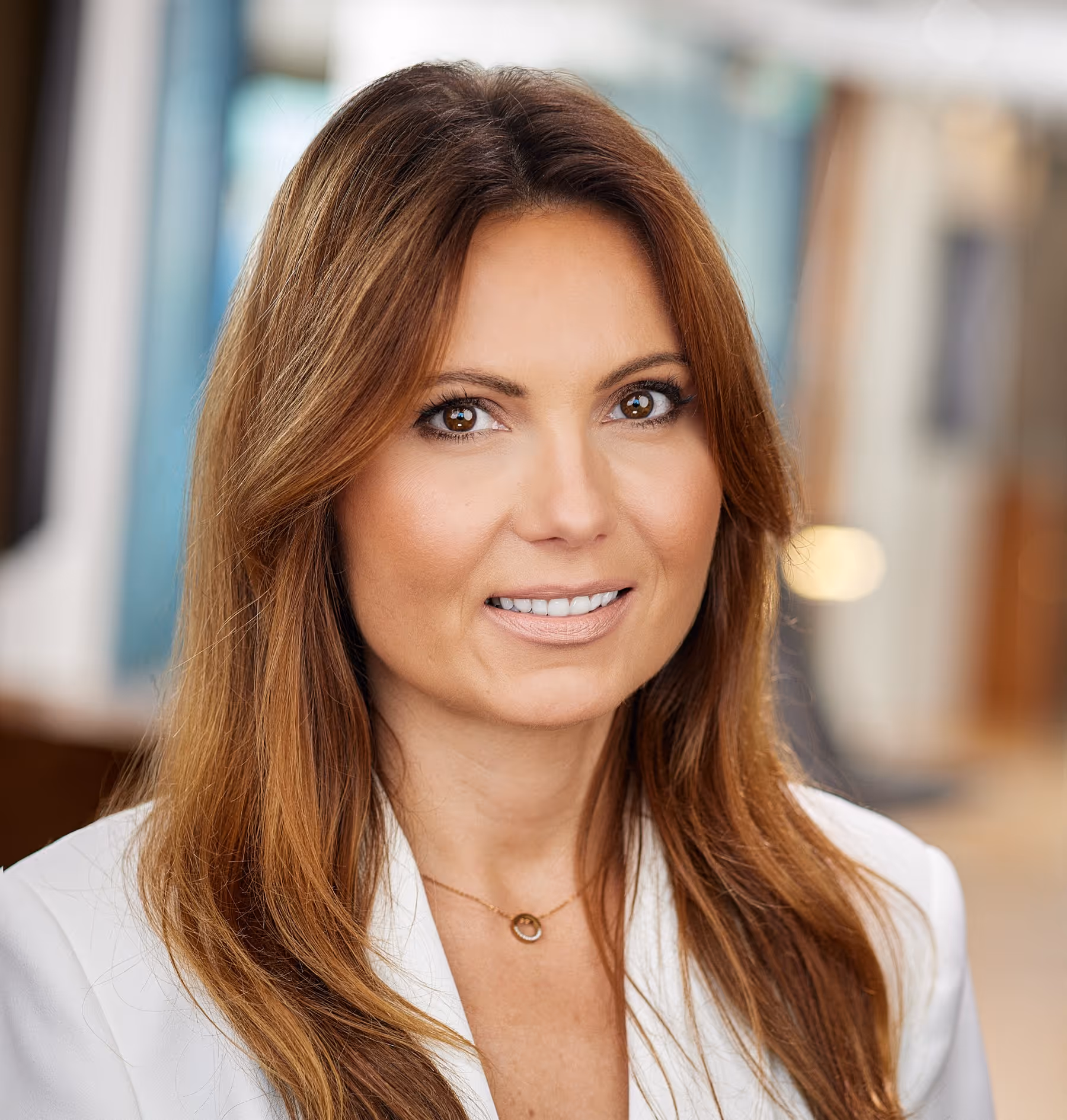 Close-up of a smiling woman with long brown hair wearing a white blazer and a delicate necklace in a softly blurred indoor setting.