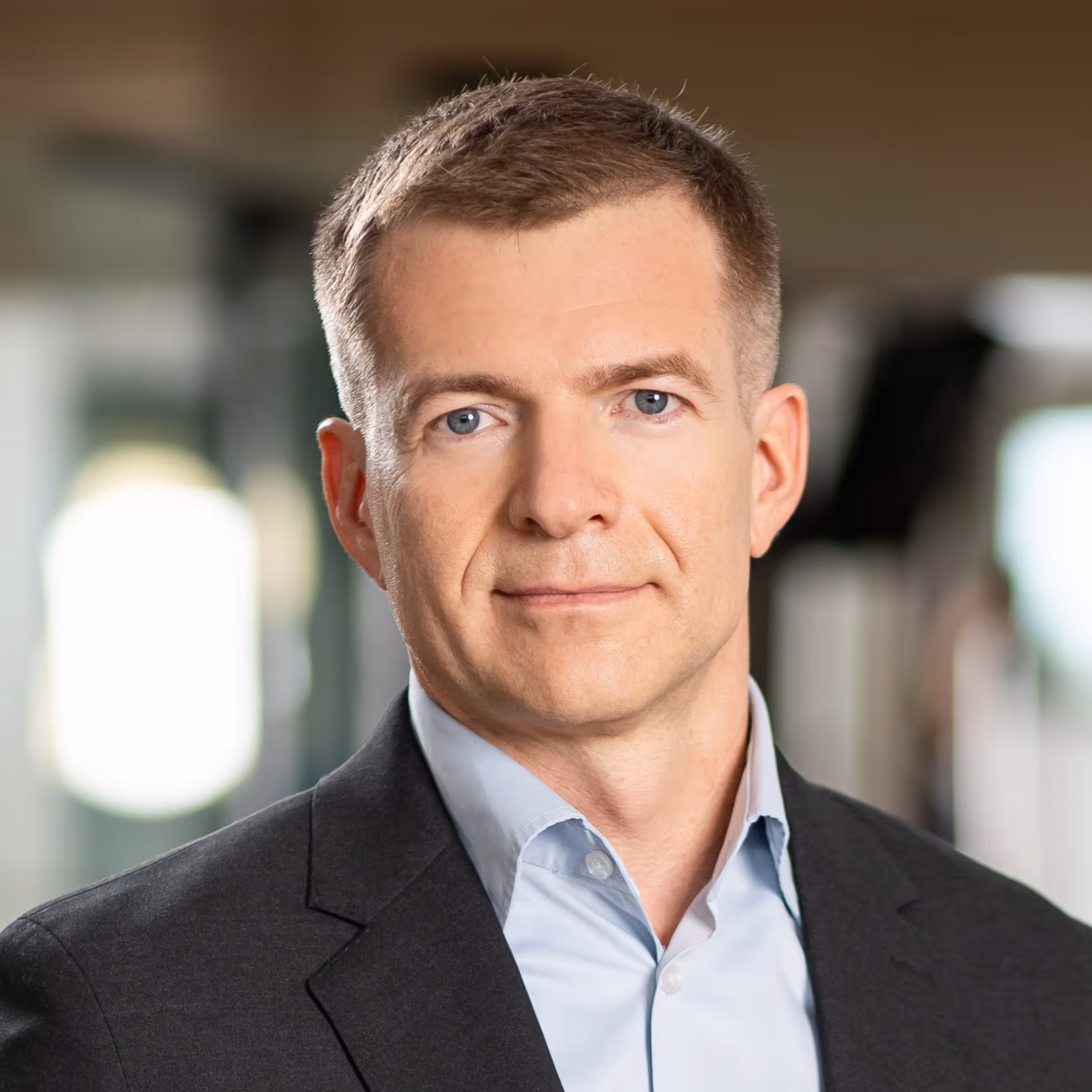 Portrait of a man with short light brown hair wearing a black blazer and light blue shirt, smiling slightly with a blurred indoor background.