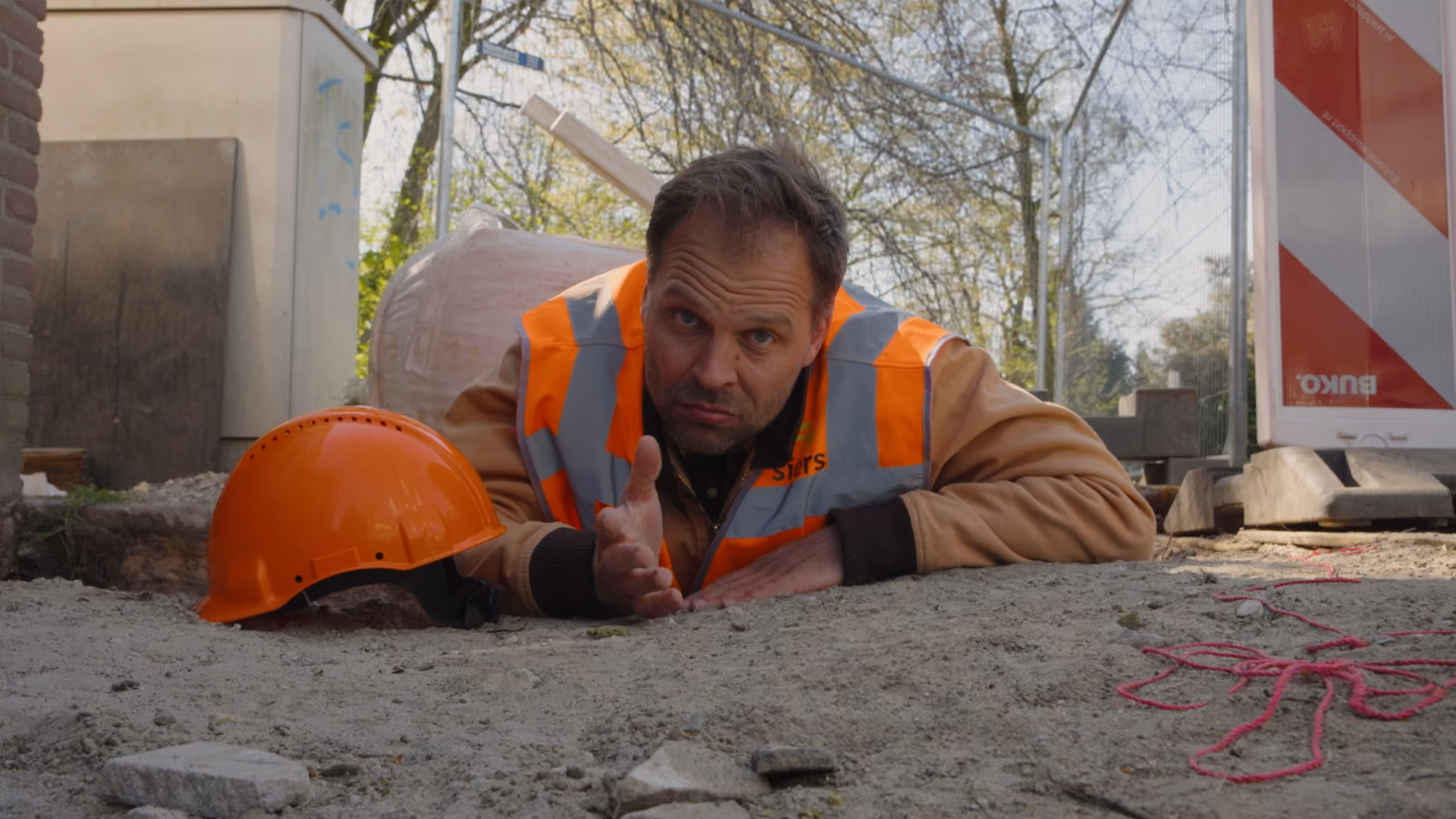 Bouwvakker met oranje hesje en helm ligt op de grond en steekt hand uit op bouwplaats.
