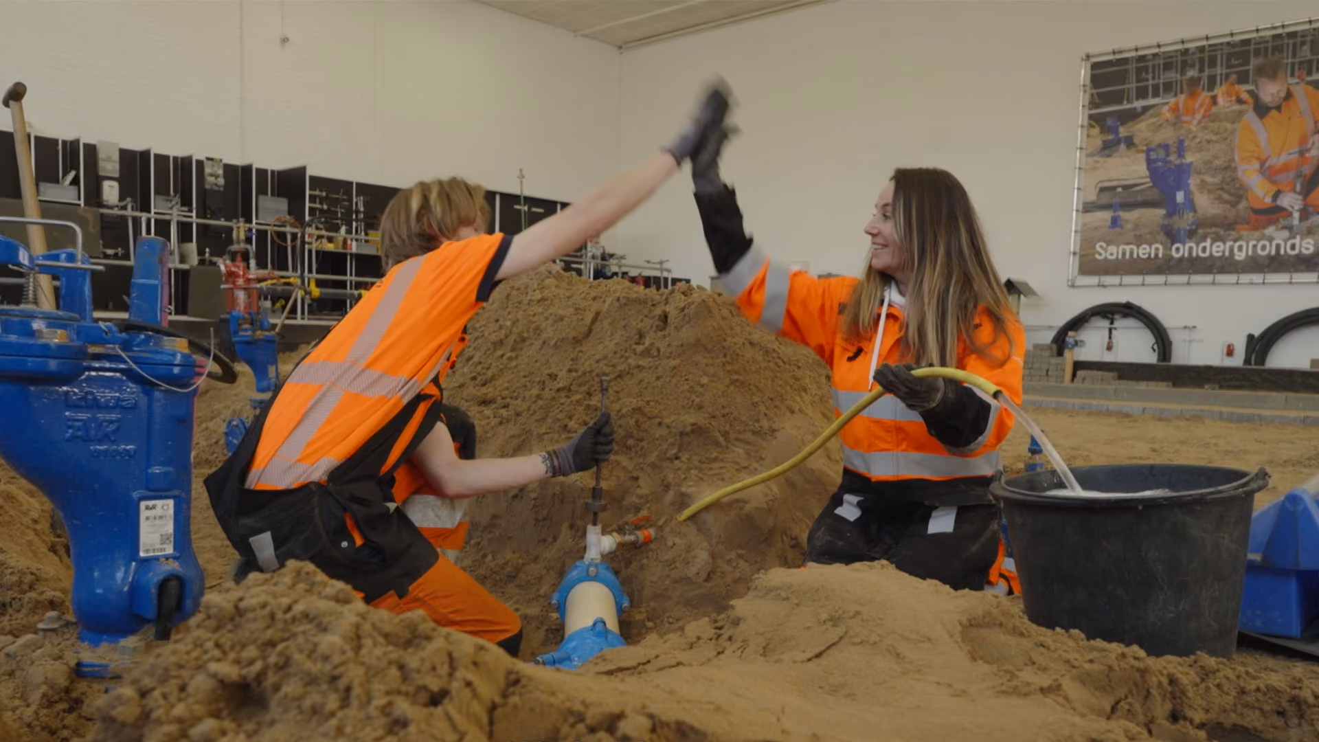 Twee bouwvakkers in oranje werkkleding en handschoenen geven elkaar een high five in een zandbak.