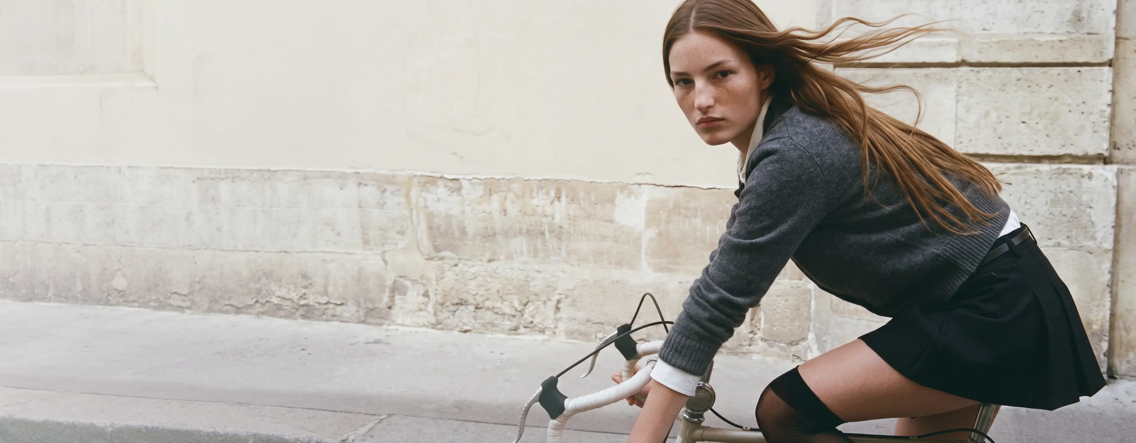 AI generated model with long auburn hair riding a bicycle past a cream stone wall