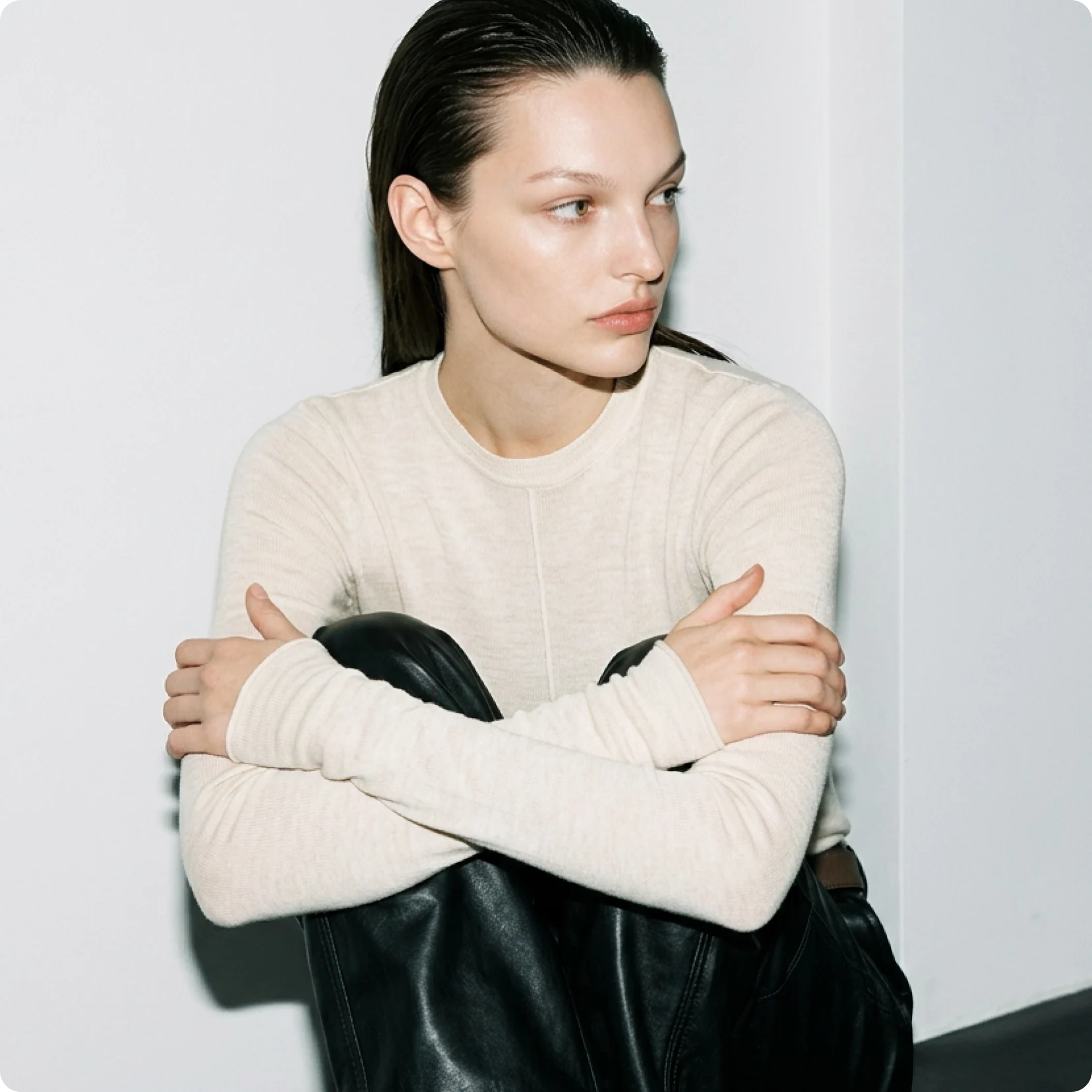 AI generated model with slicked-back hair in a cream knit top and black leather trousers, seated against a white wall