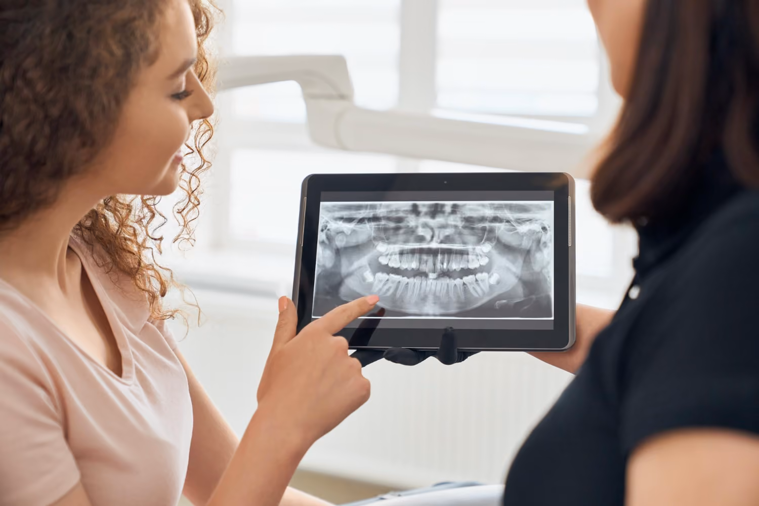 Woman pointing at dental X-ray shown on tablet while talking to a dentist in clinic.