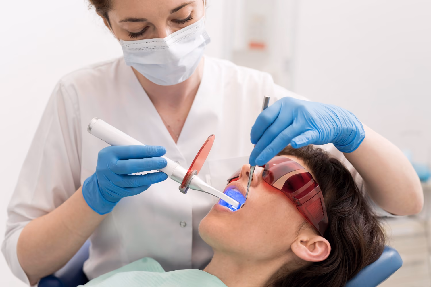 Dentist in mask and gloves using a curing light on a patient's teeth wearing protective glasses.