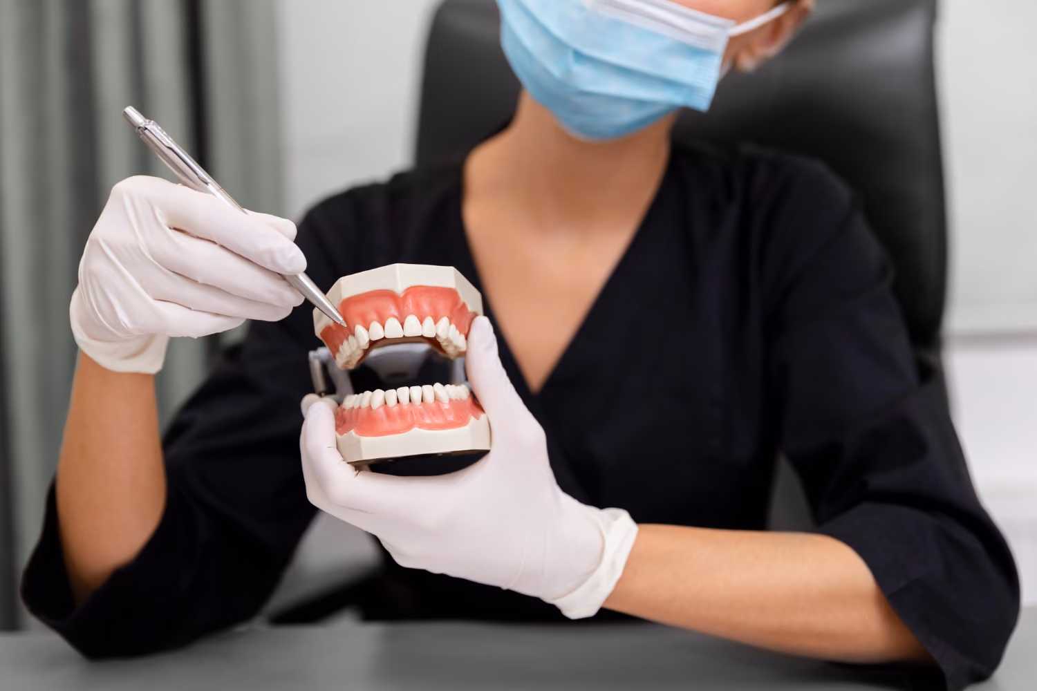 Dentist wearing gloves and mask holding a dental model and pointing at teeth with a pen.