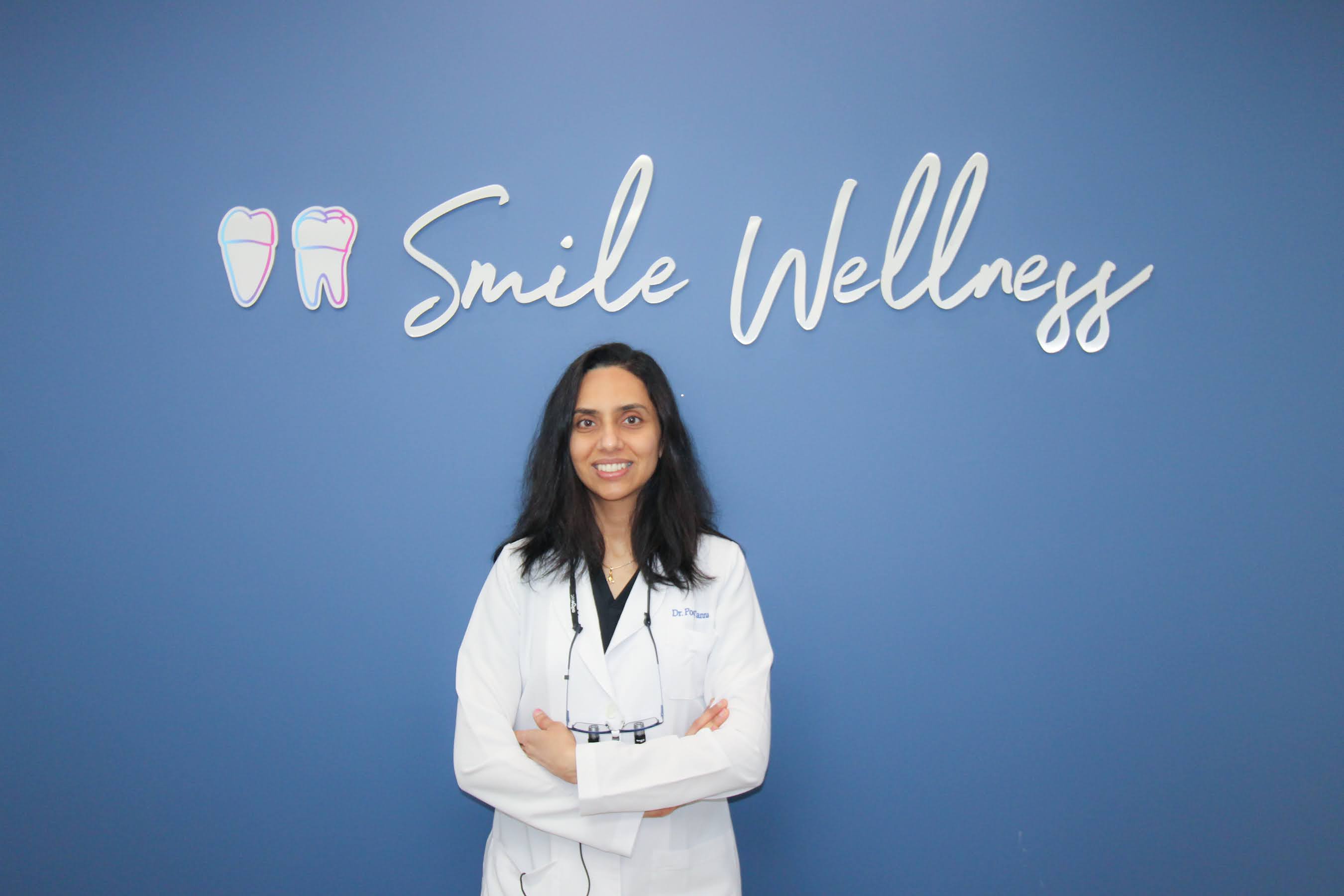Female dentist in white lab coat smiling and standing with arms crossed in front of Smile Wellness sign.