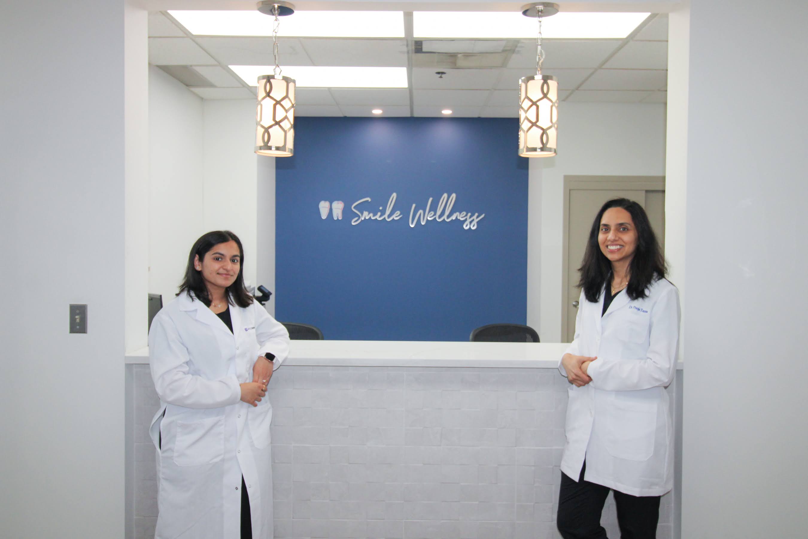 Two female dentists in white coats standing by a reception desk with a Smile Wellness sign on blue wall.