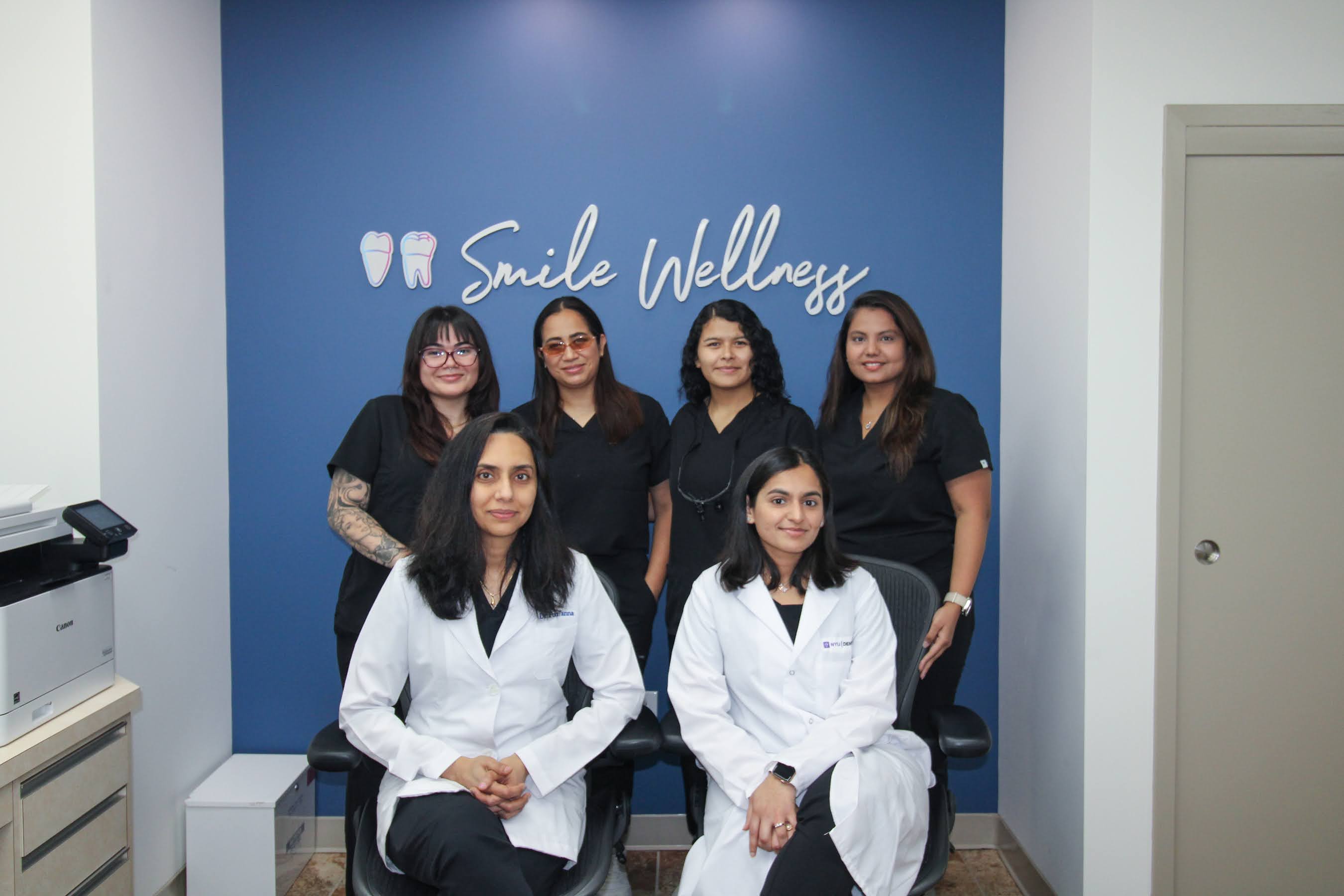 Six dental professionals posing inside Smile Wellness office, two in white coats seated, four in black scrubs standing.