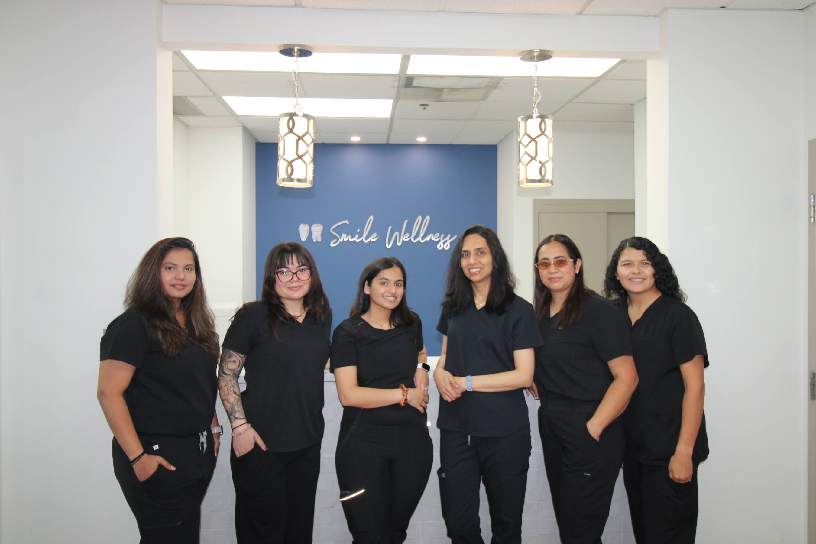 Six women in black scrubs stand and smile in front of a blue 'Smile Wellness' sign indoors.