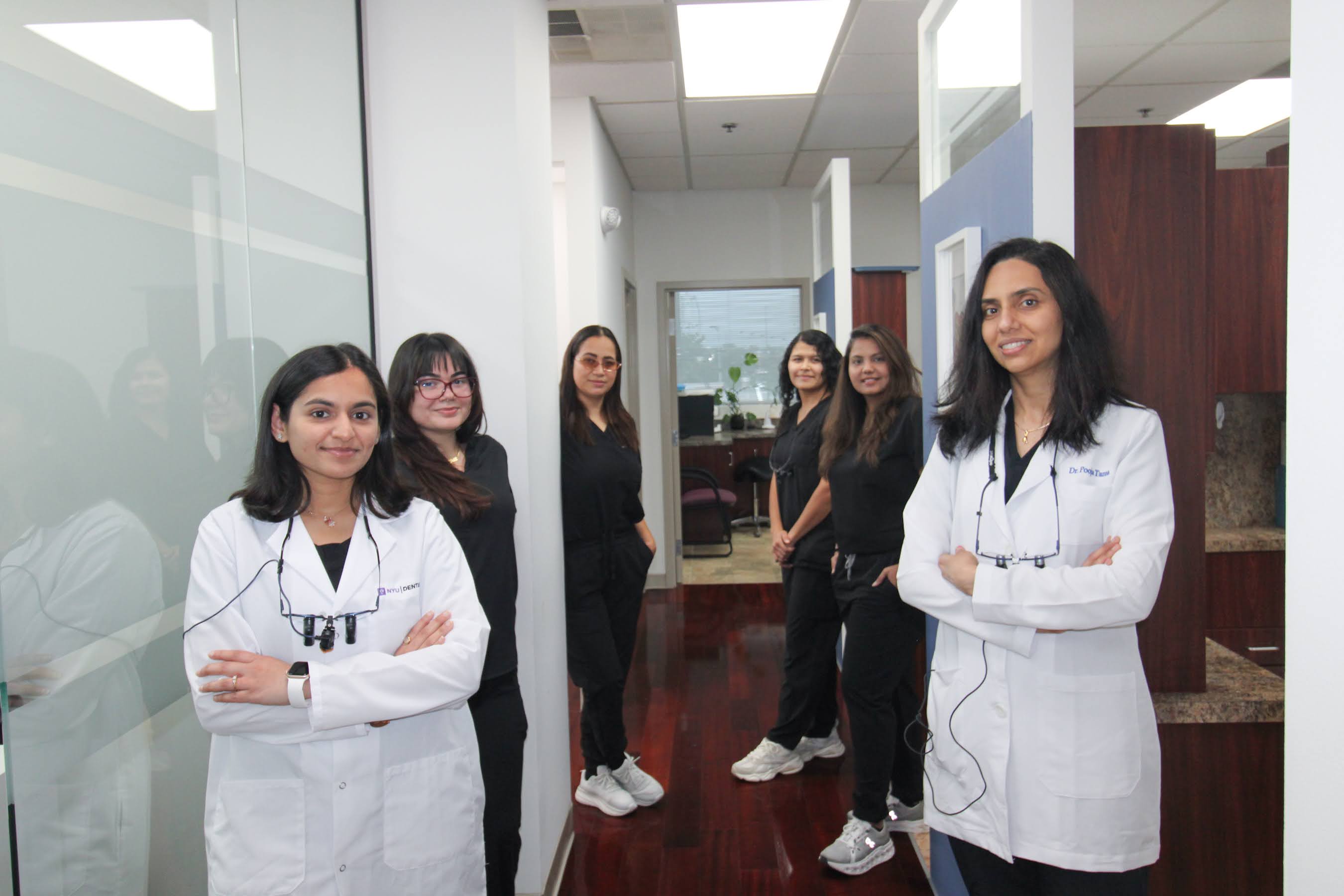 Six female medical professionals standing in a clinic hallway, two wearing white coats and four in black scrubs.