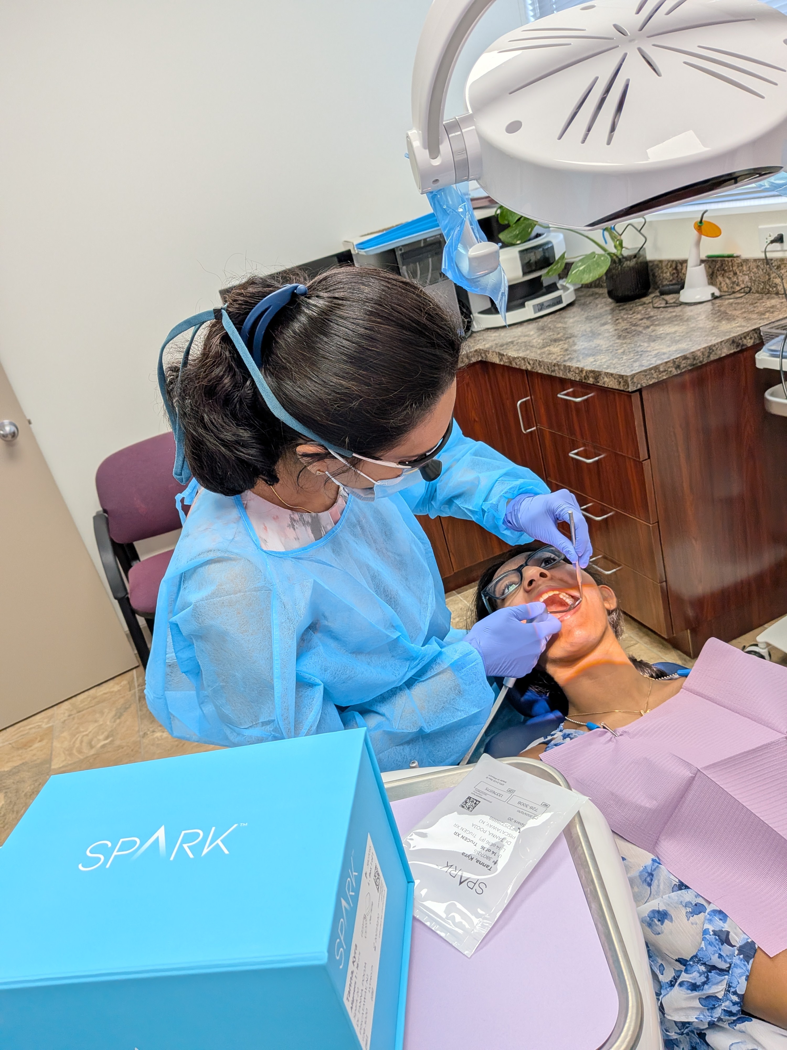 Dentist in blue gown treating patient wearing glasses in dental chair under exam light.