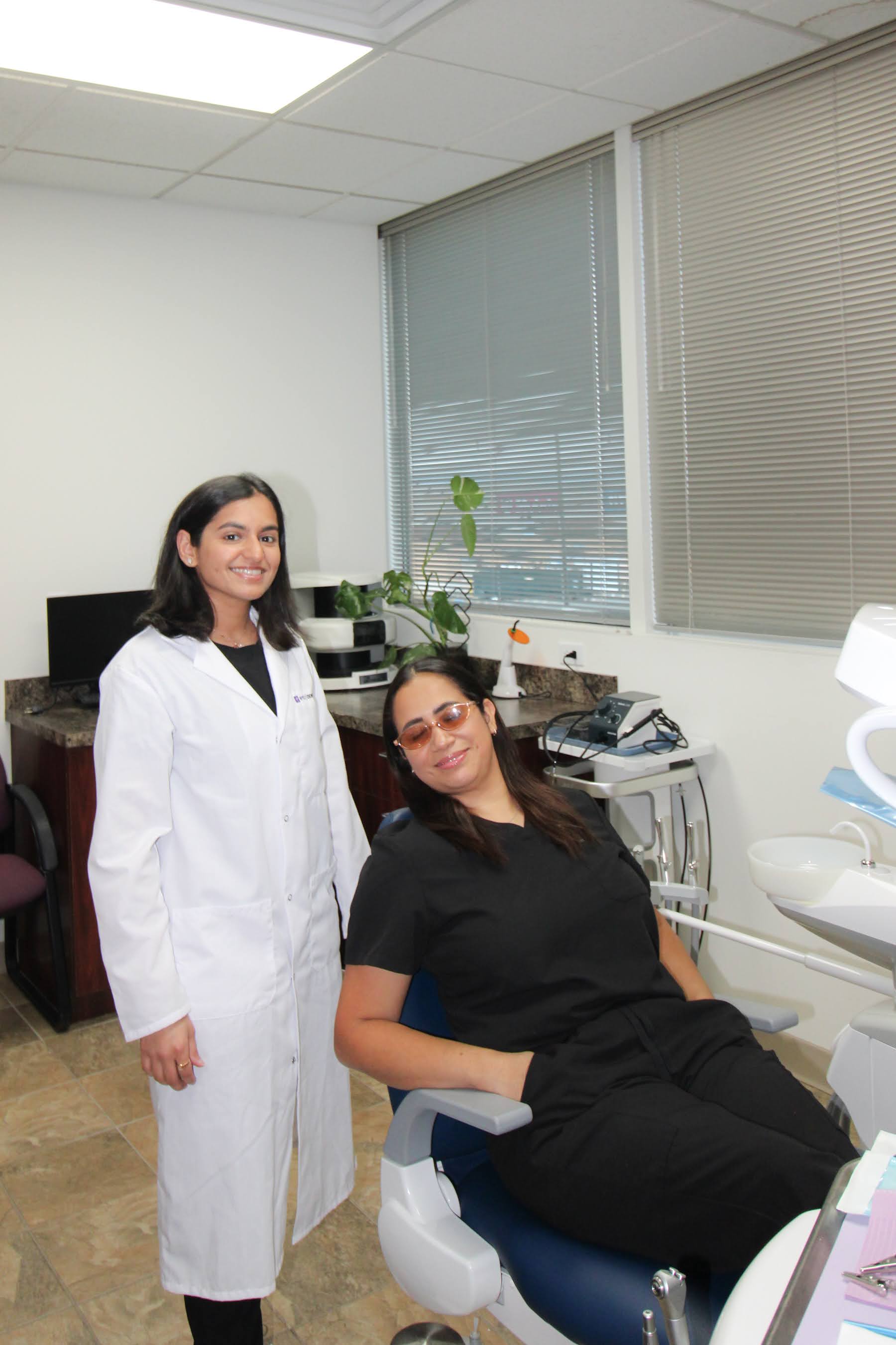 Smiling female dentist in white coat with a relaxed patient seated in dental chair wearing protective glasses.