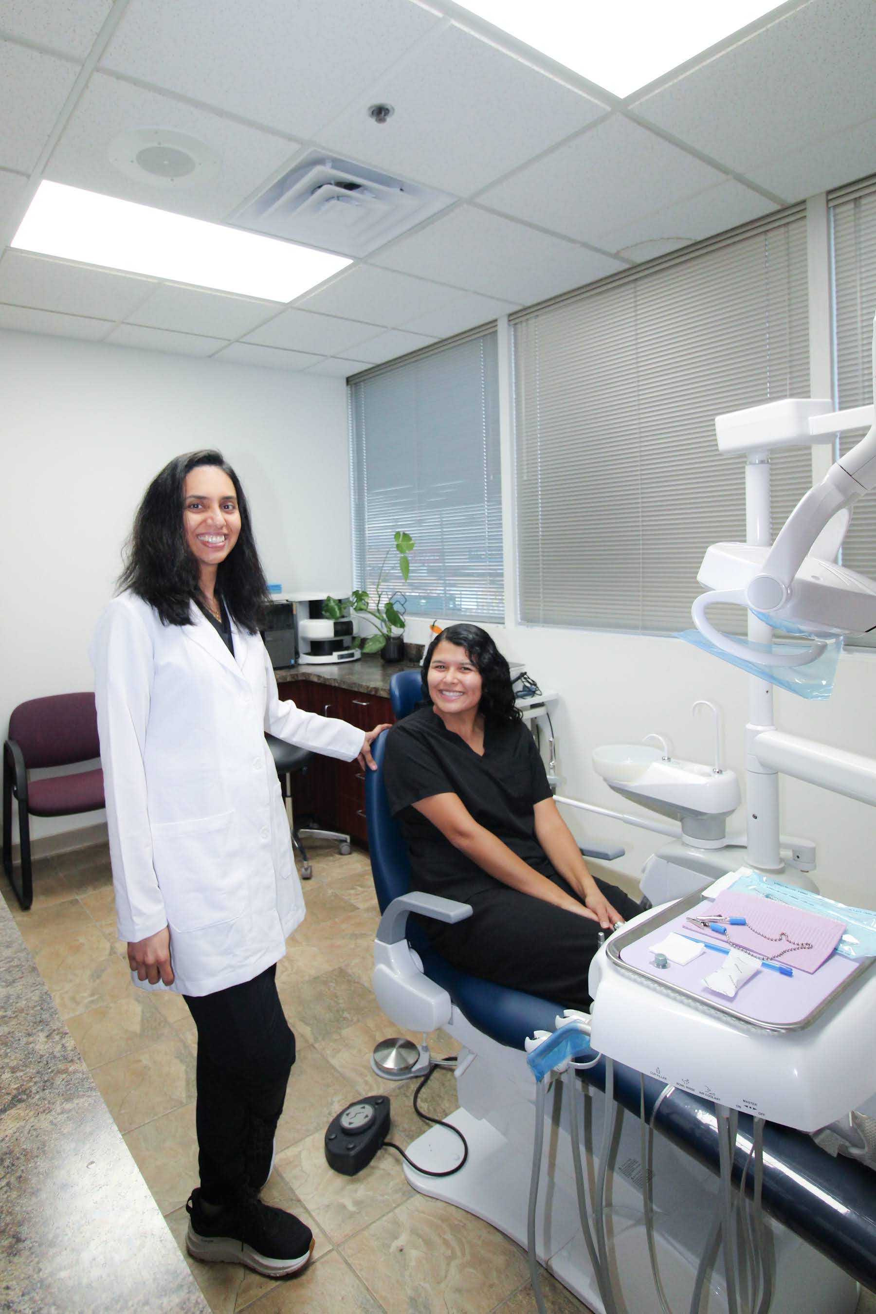 Smiling dentist in white coat stands beside a seated patient in black scrubs in a dental office.