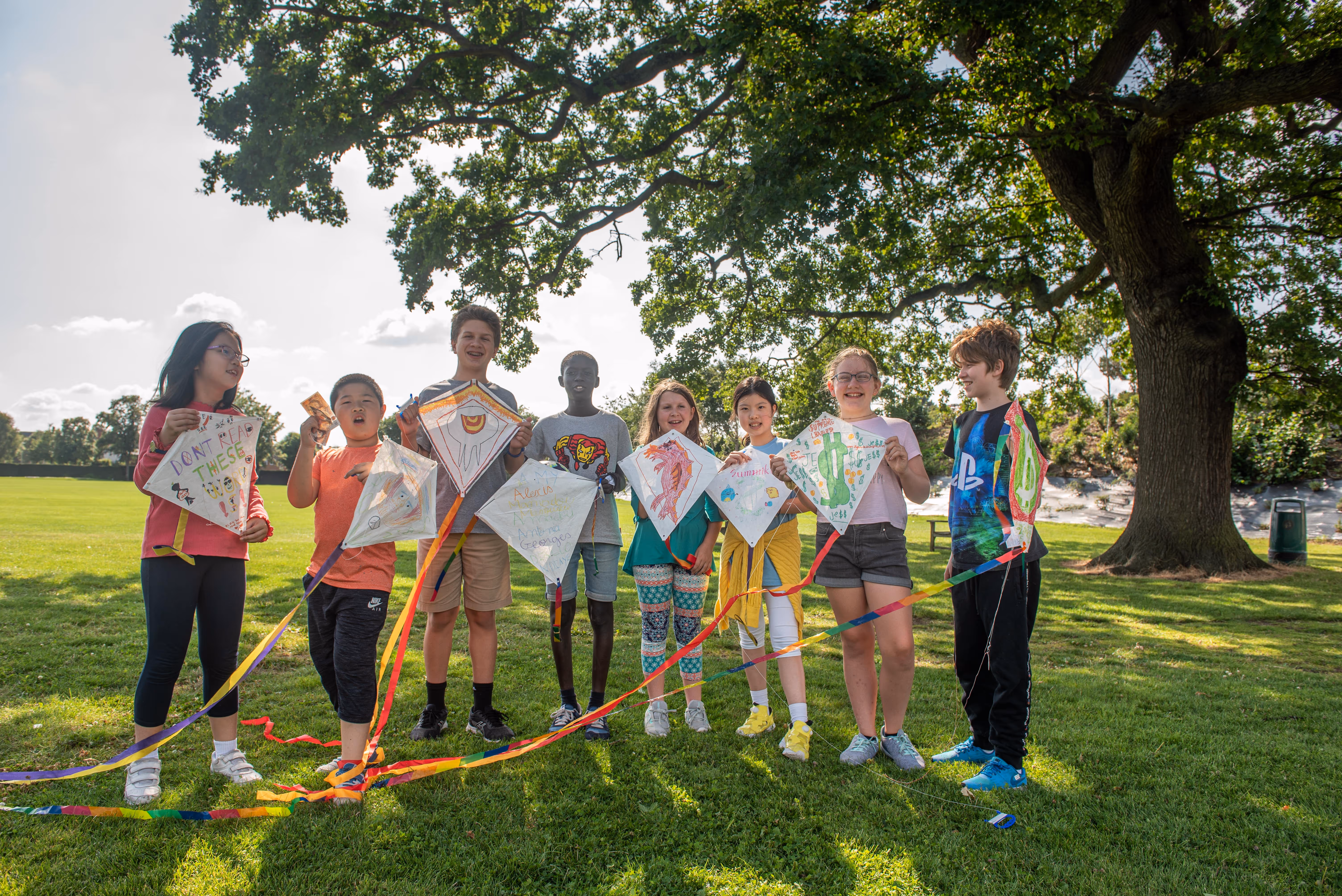 Group of children standing on grass holding decorated kites under a large tree