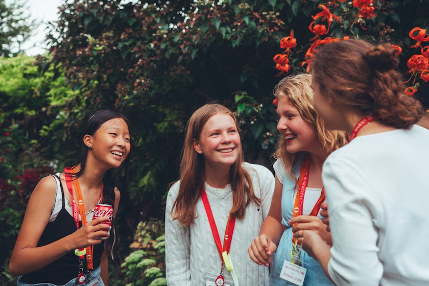 Four teenage girls smiling and chatting outdoors with greenery and orange flowers in the background.