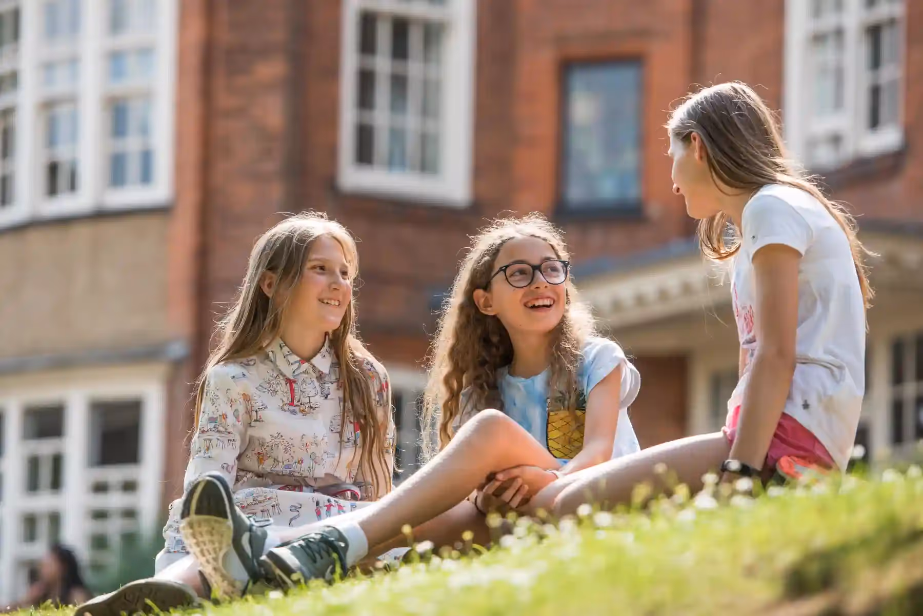 Three girls sitting on grass outside a brick building, smiling and talking together.