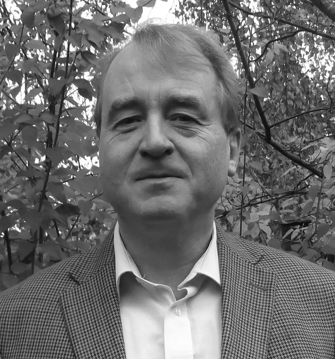 Black and white portrait of a middle-aged man wearing a checkered jacket and white shirt, standing outdoors with foliage in the background.