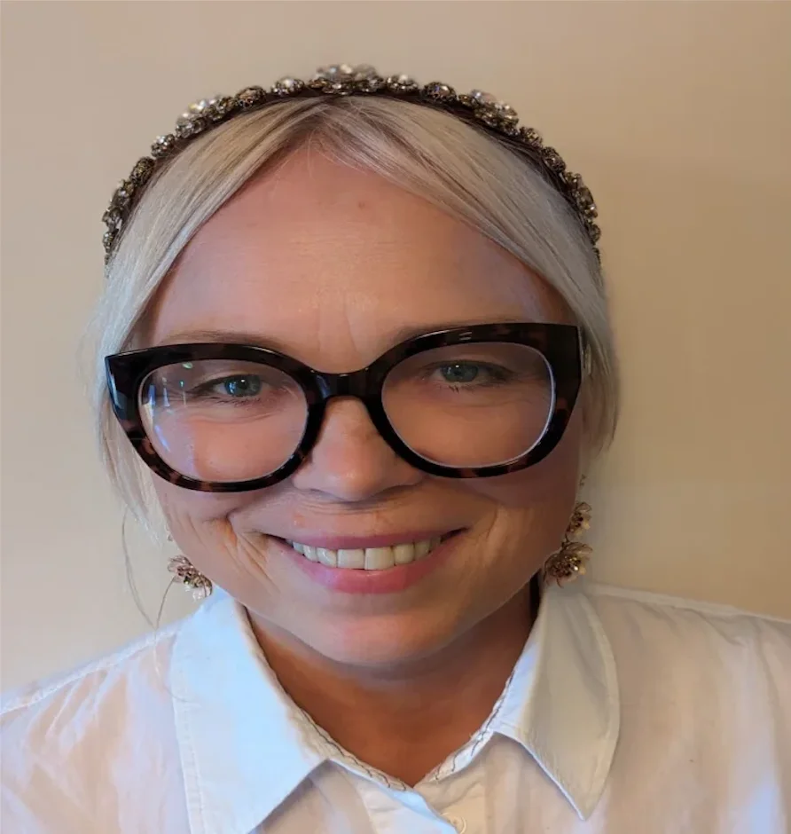 Smiling woman with light blonde hair, wearing dark-framed glasses, a jeweled headband, and a white shirt against a plain background.