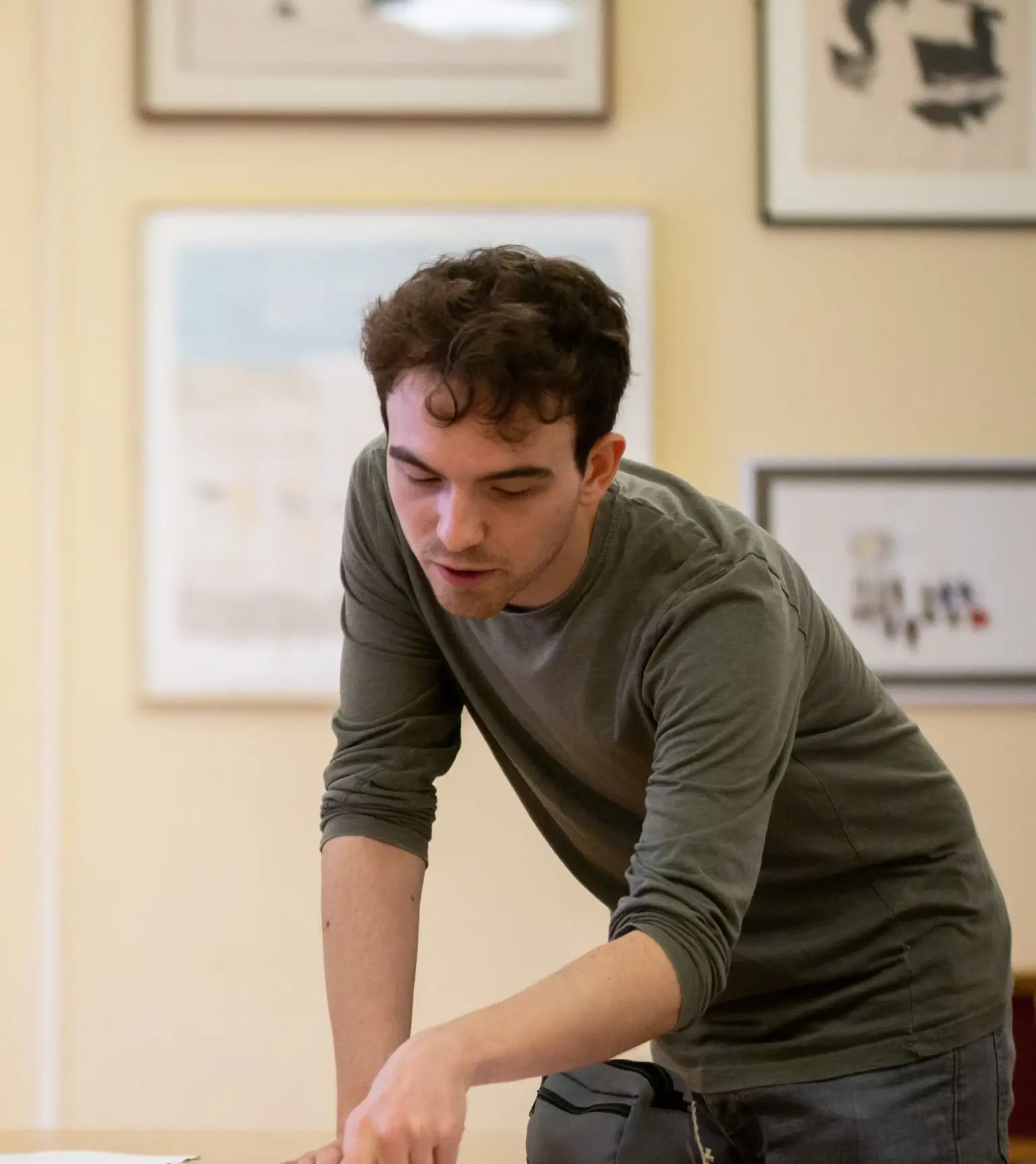 Young man with curly brown hair leaning over a table, pointing at something out of view in a room with framed pictures on the wall.