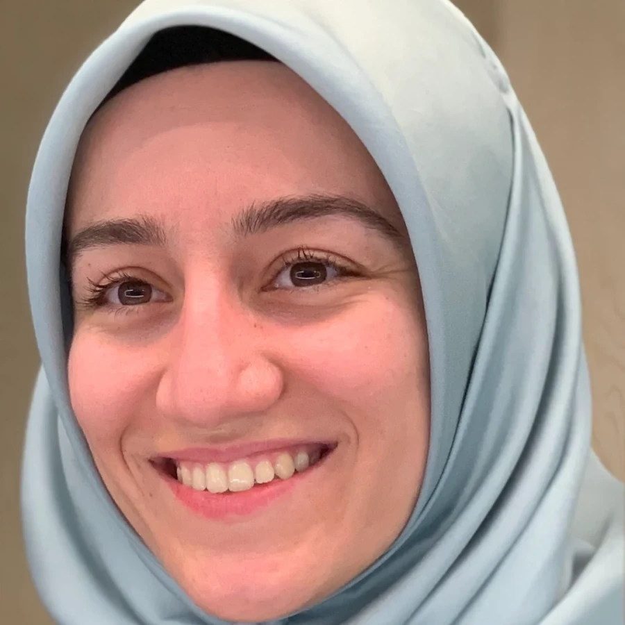 Close-up of a smiling woman wearing a light blue hijab with a neutral background.