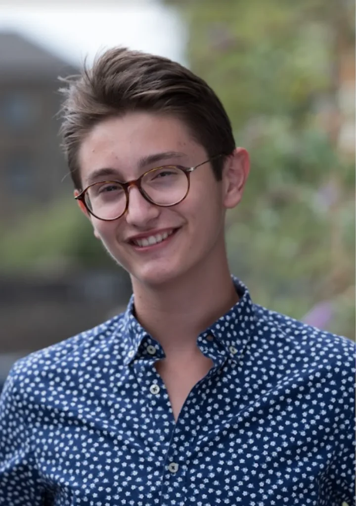 Young person with short brown hair and glasses smiling, wearing a blue shirt with white floral pattern.