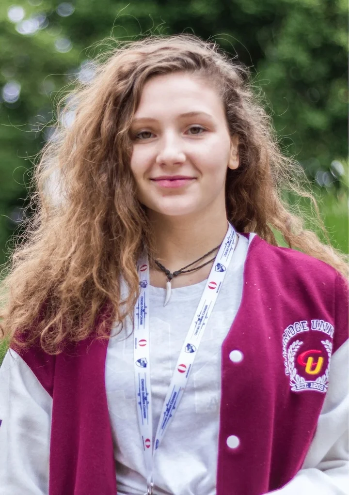 Young woman with long curly hair wearing a maroon and white varsity jacket and a lanyard, smiling outdoors.