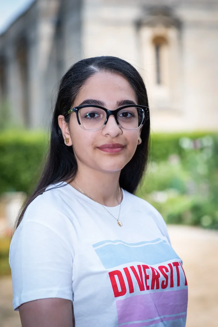 Young woman wearing glasses and a white T-shirt with the word 'DIVERSITY' in red letters, outdoors with blurred greenery and a building in the background.