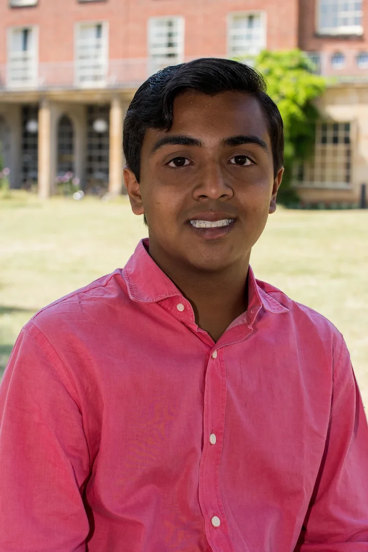 Young man wearing a bright pink button-up shirt smiling outdoors in front of a building with windows and columns.