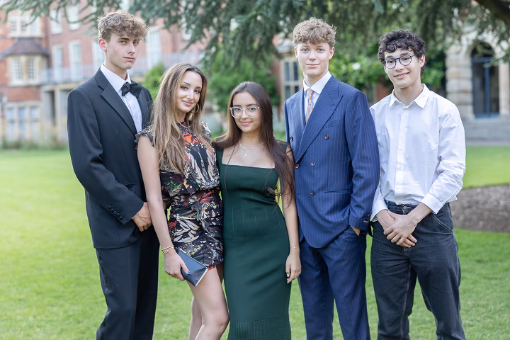 Five young adults dressed formally, three men in suits and two women in dresses, standing outdoors on green grass in front of a brick building with trees.