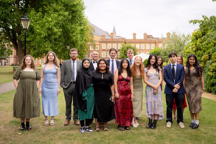 Group of diverse young adults dressed in formal and semi-formal attire standing on grass in front of a historic brick building with trees around.