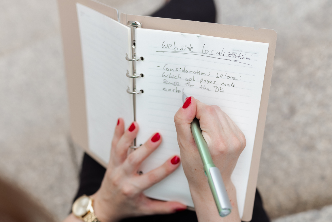 Person with red nail polish writing notes on website localization in a notebook.