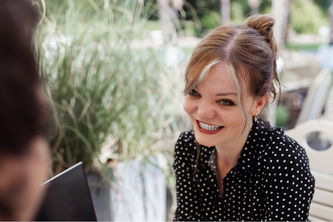 Smiling woman with polka dot blouse sitting outdoors, looking at a laptop.