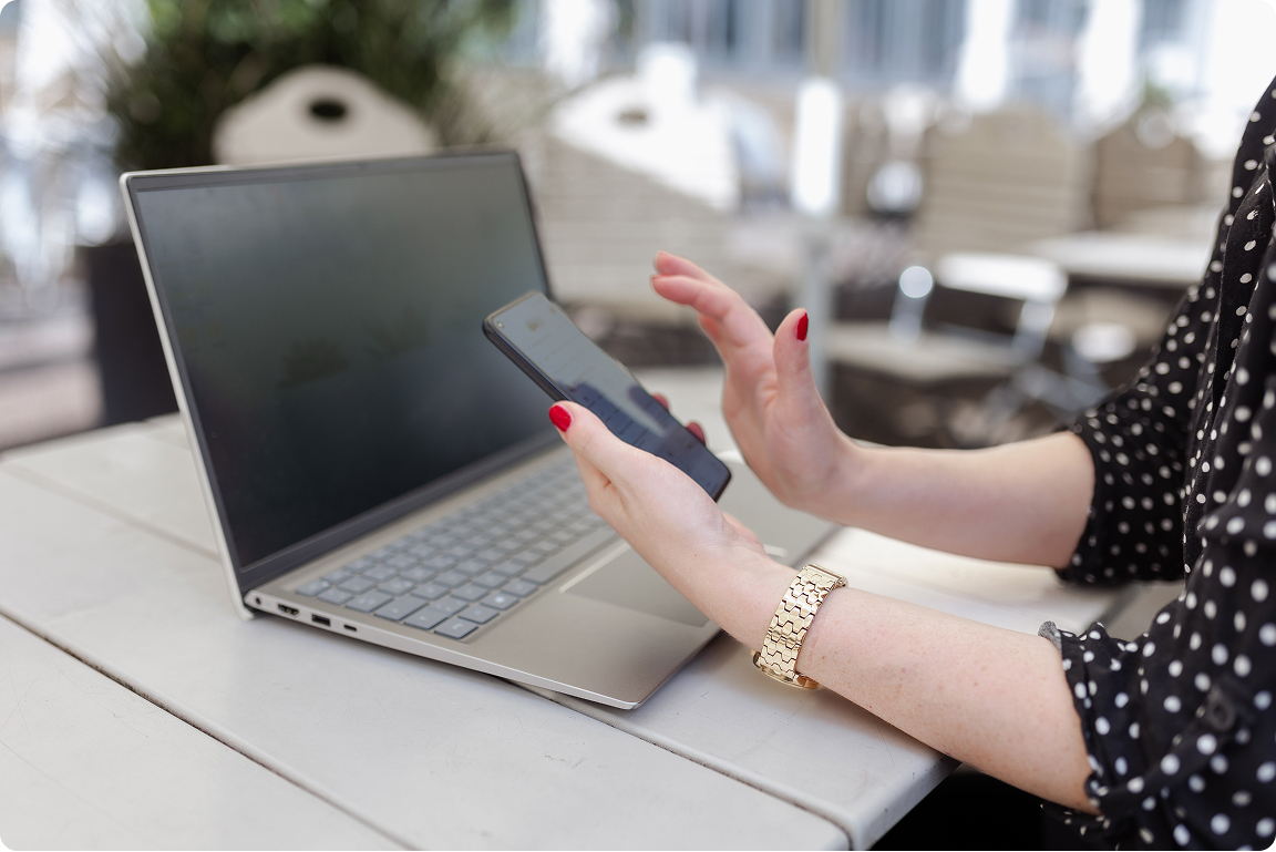 Person with red nail polish and gold watch using smartphone next to a laptop on a table outdoors.