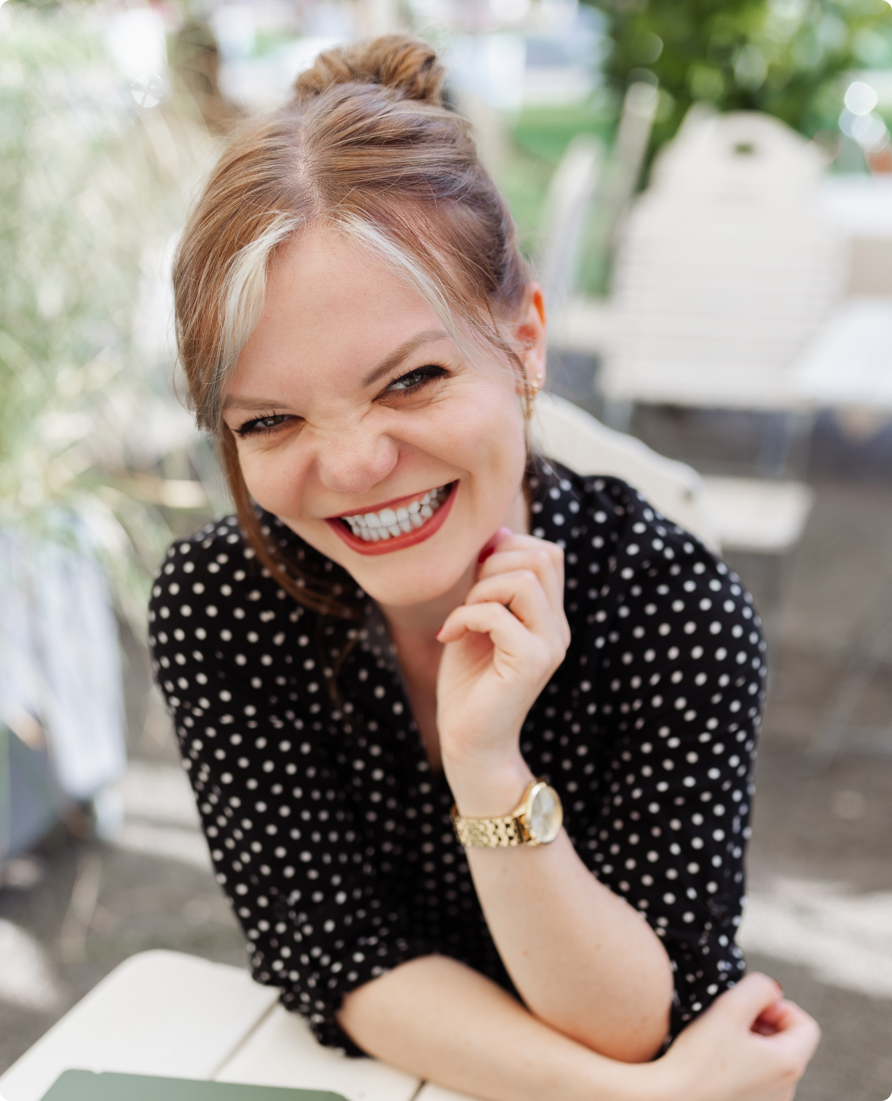 Smiling woman in a black polka dot shirt with a gold wristwatch, sitting at an outdoor table.