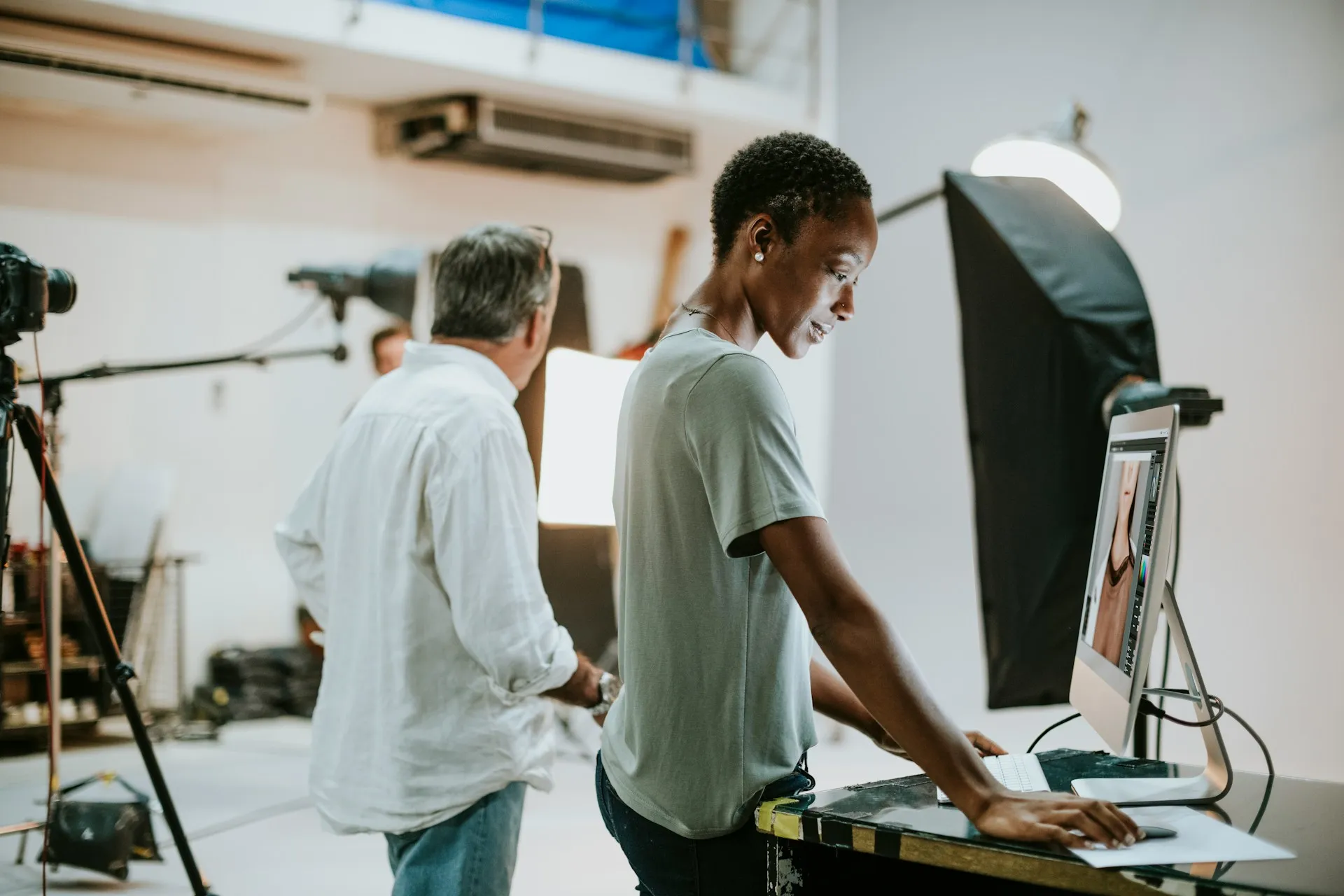 Two people in a photography studio reviewing photos on a desktop computer, surrounded by studio lighting and camera equipment.