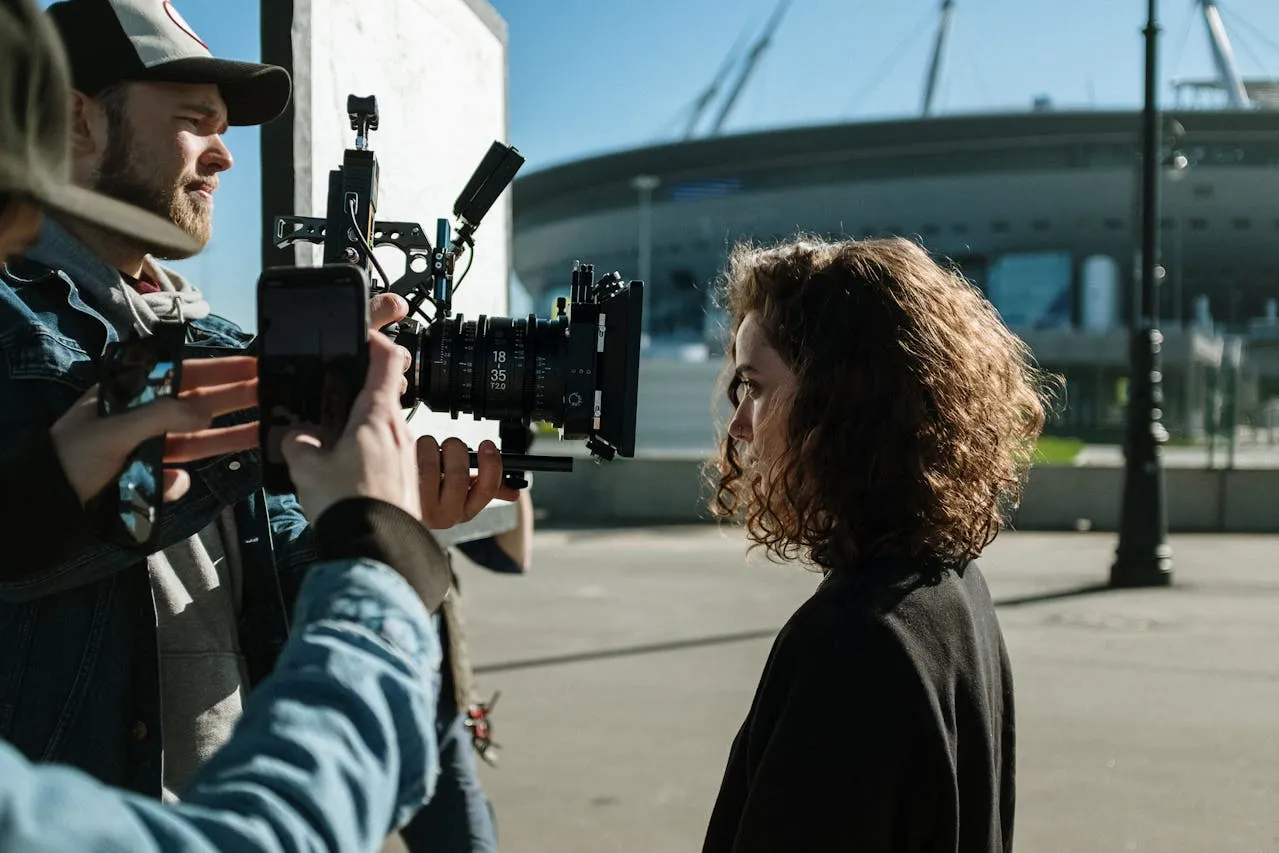 Filmmaker with a camera rig filming a woman with curly hair outdoors near a stadium.