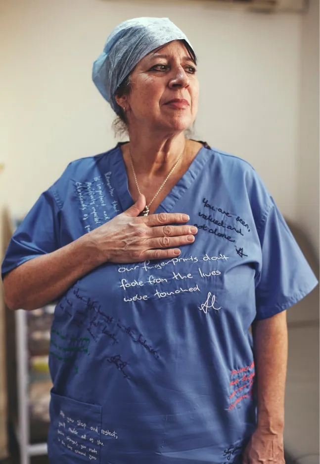 Full-length portrait of a woman in blue scrubs standing with one hand on her chest, featuring embroidered and handwritten statements across the uniform.