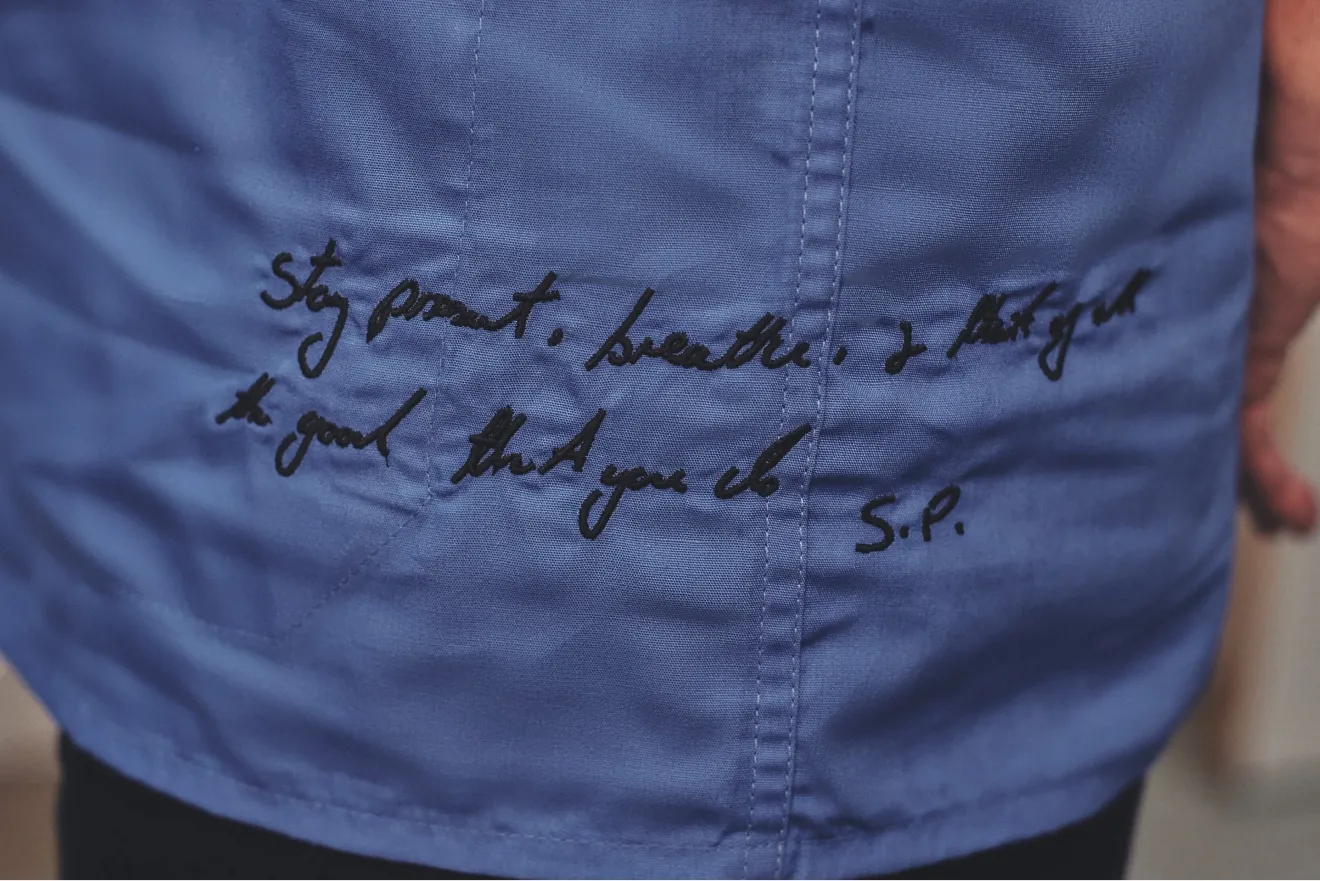 Close-up of blue scrubs with handwritten black text reading, “Stay present, breathe, I ask you to trust that you do.”