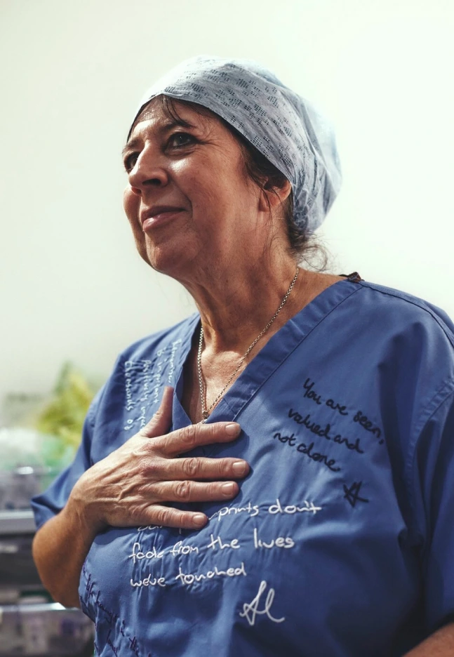 Portrait of a woman in blue scrubs and surgical cap with one hand on her chest, surrounded by handwritten and embroidered messages.