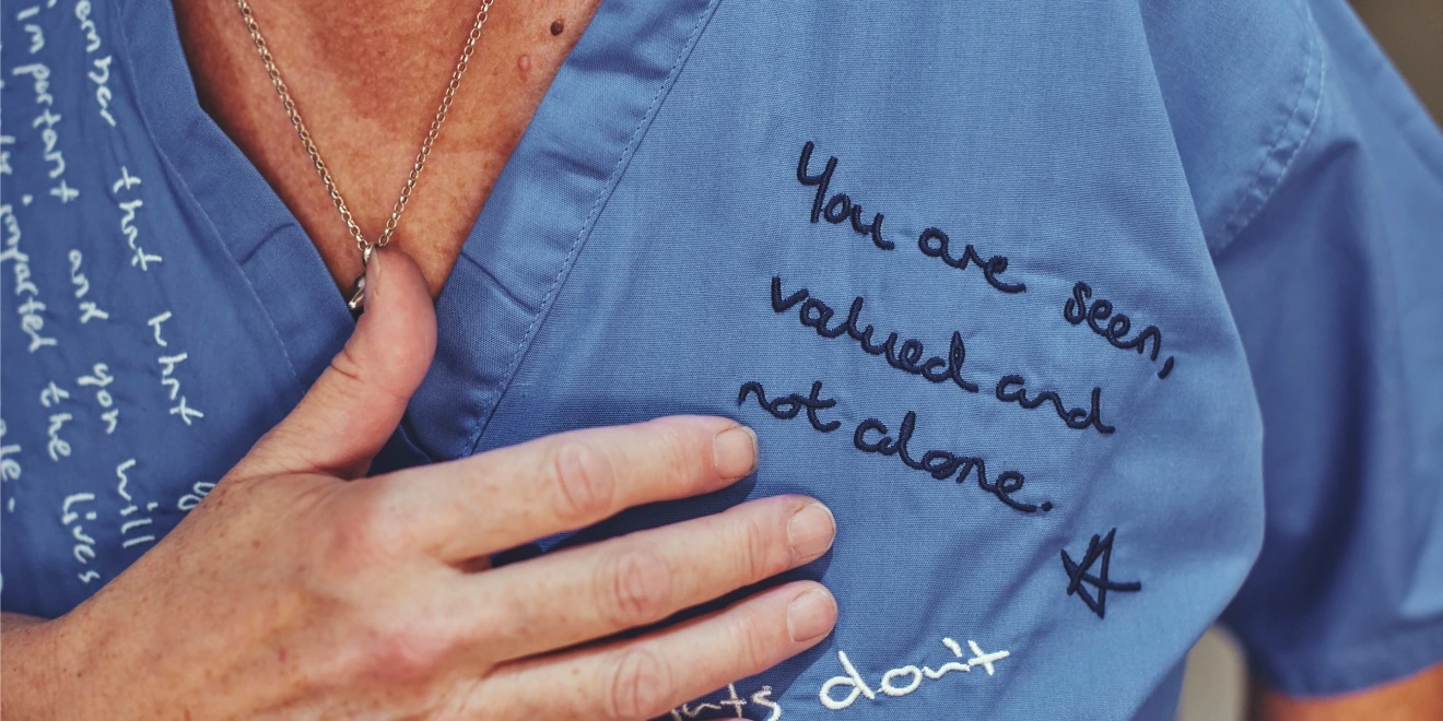 Close-up of the chest area of blue scrubs showing the handwritten message, “You are seen, valued and not alone,” beside the woman’s hand.