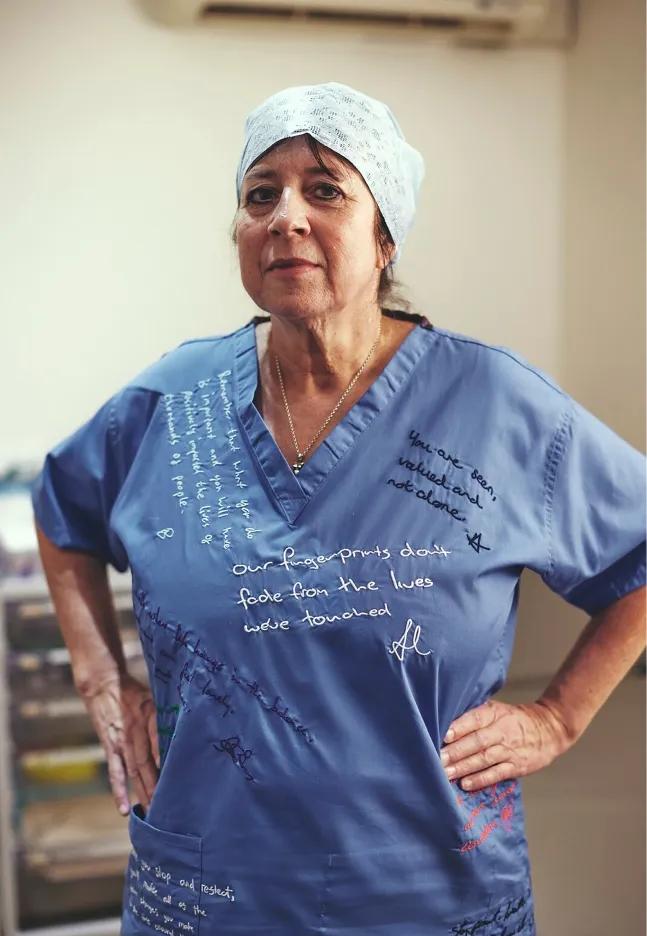Full-length portrait of a woman in blue scrubs standing with hands on hips, her uniform covered in supportive and reflective stitched messages.
