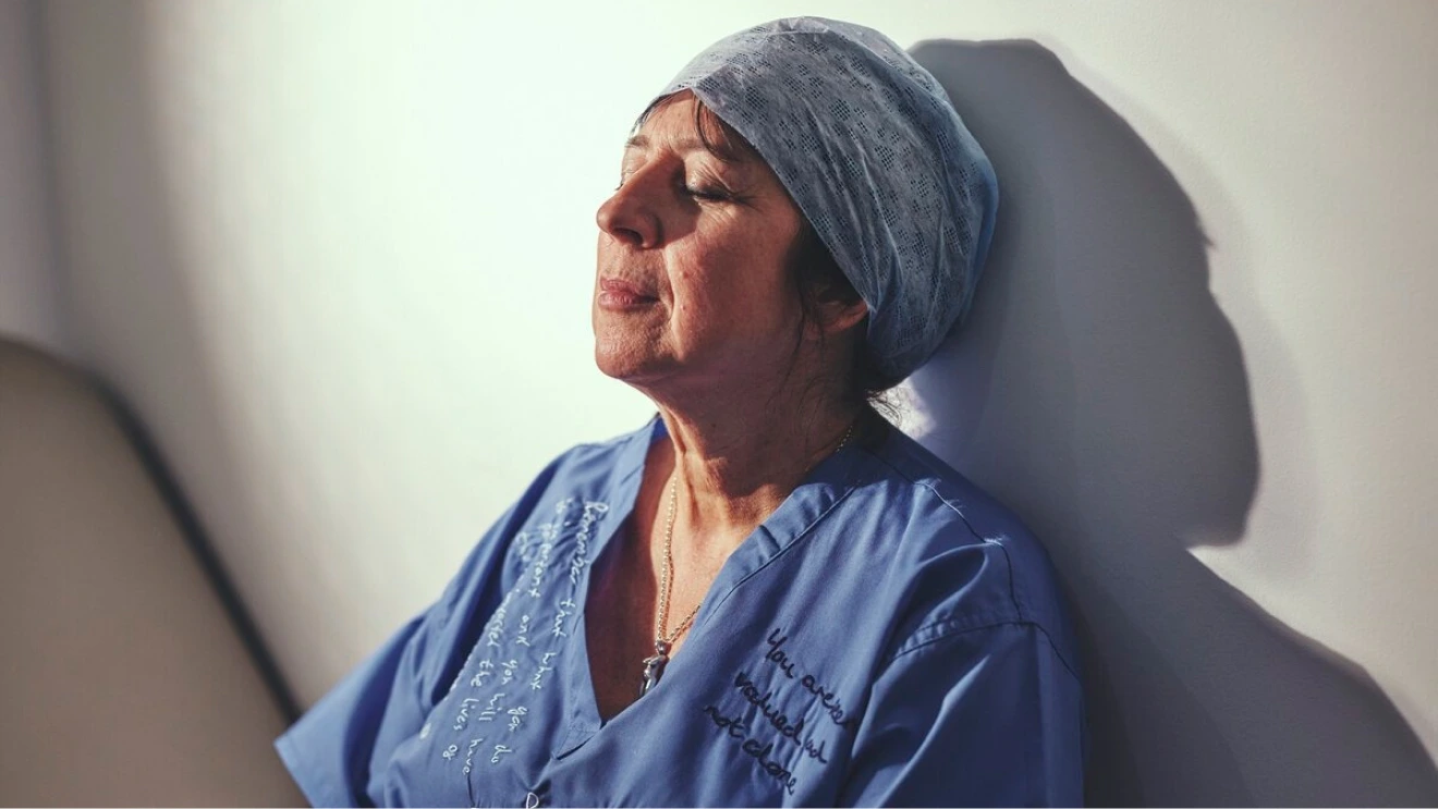 Portrait of a woman in blue scrubs leaning back against a wall with eyes closed, lit by strong side light and shadow.