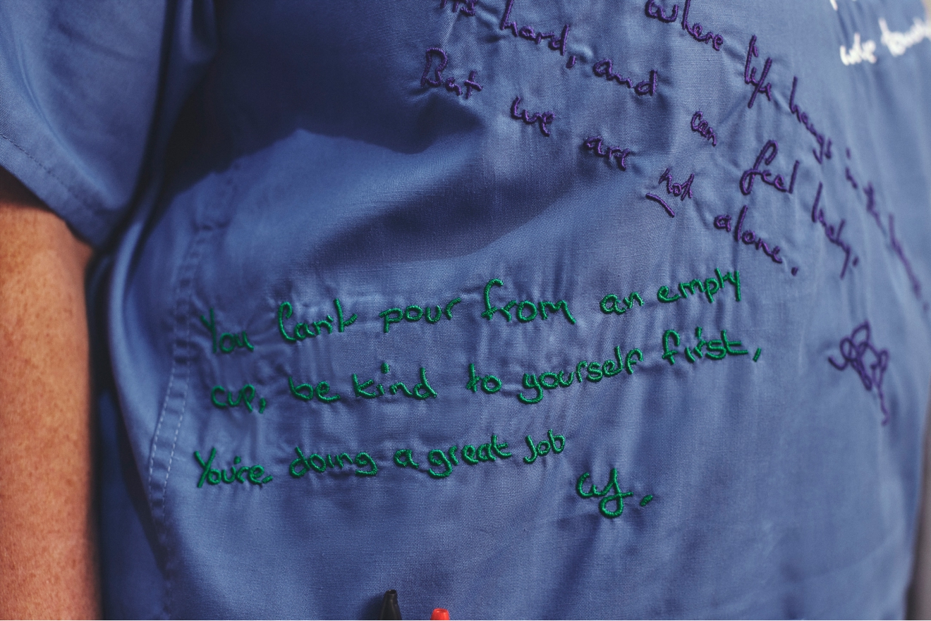 Close-up of blue scrubs covered in handwritten messages, including green text about self-care and doing a great job.