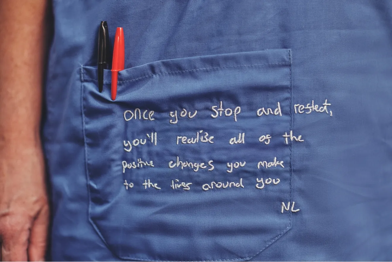Close-up of the pocket area of blue scrubs with a red and a black pen tucked inside, embroidered with the message: “Once you stop and rest, you’ll realise all of the positive changes you make to the lives around you.