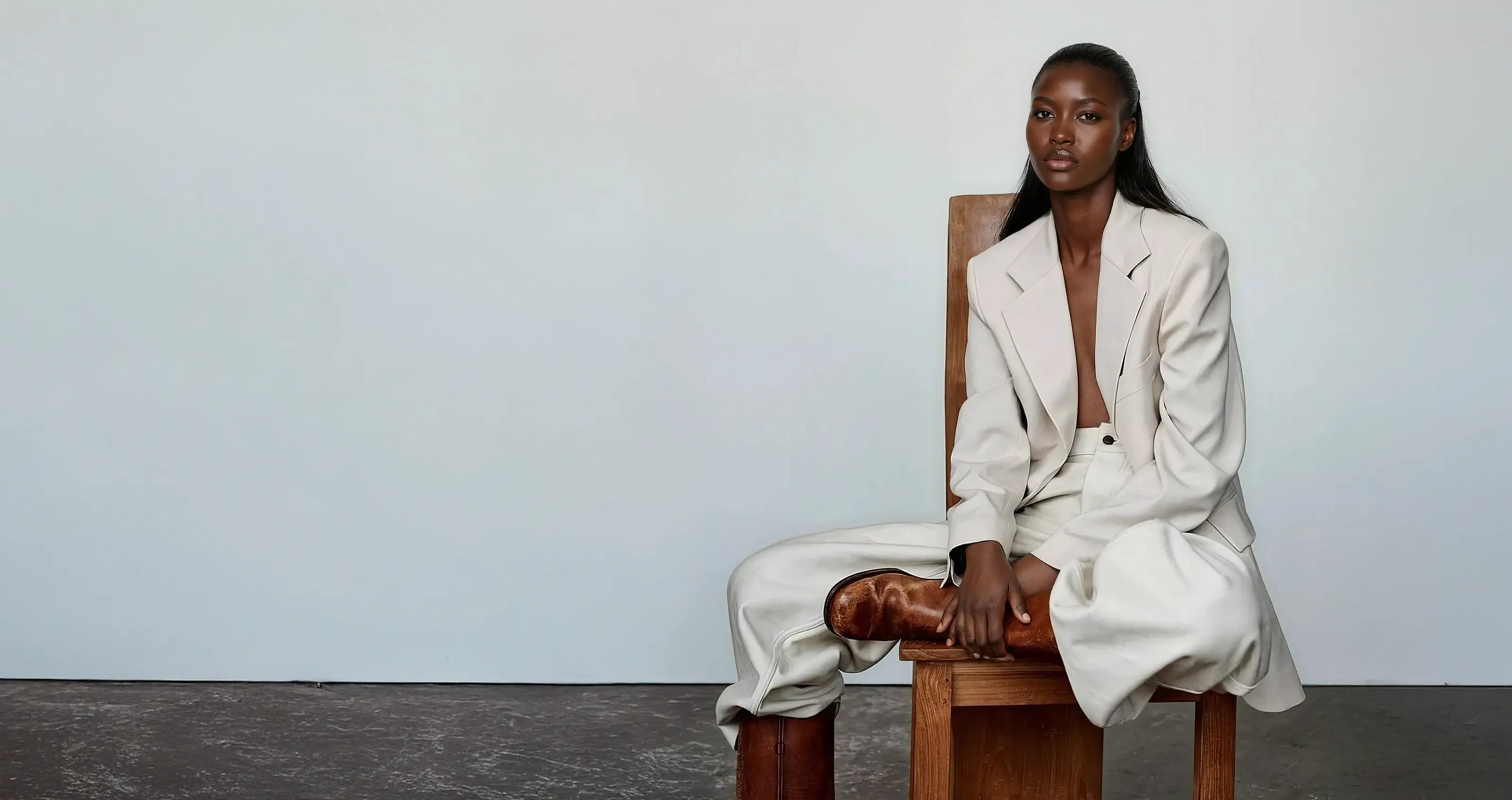 Photo of black female AI fashion model wearing a white suit and light brown boots sitting on a chair.