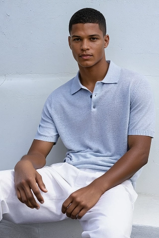 A black male AI fashion model named Noah, created by Botika sitting against a wall wearing a powder blue short sleeved knit shirt and white pants. 