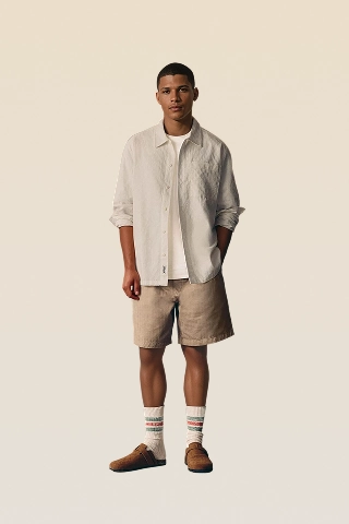 Michael, a black male AI fashion model created by Botika wearing tan shorts, a light colored long sleeve button down shirt with sleeves rolled up and a tshirt underneath. 