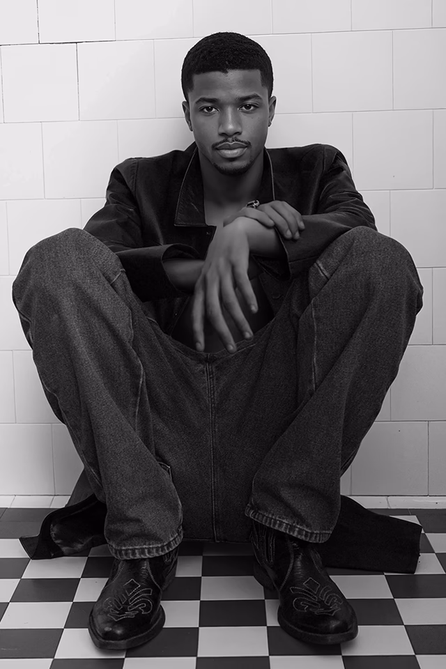 Black and white photo of Eric, a Botika created black male AI fashion model sitting on a black and white tile floor wearing dark jeans and a dark blazer.